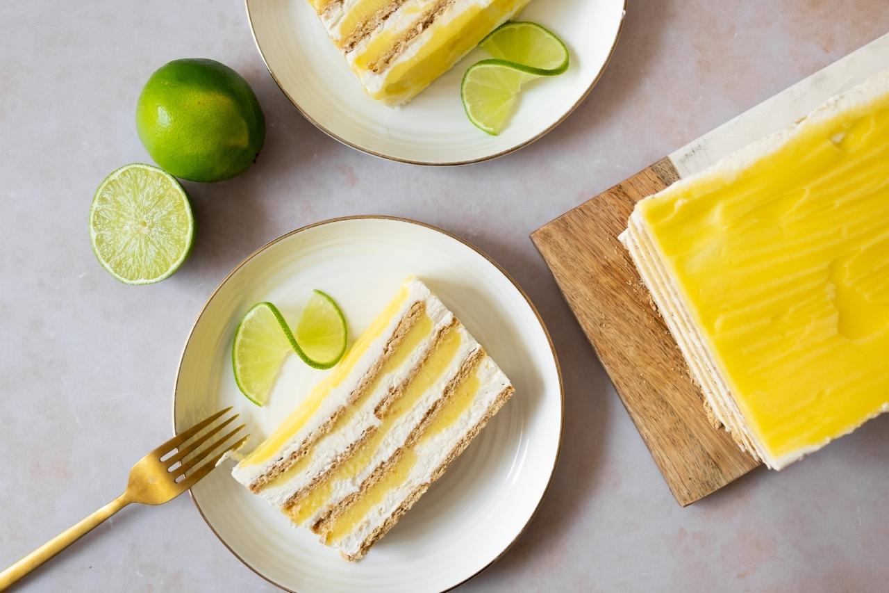 An overhead view of a slice of key lime box cake on a white plate beside the cake itself and two limes