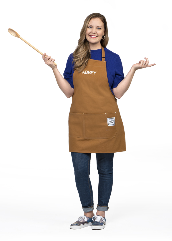 Abbey White wears jeans, a t-shirt and a brown apron while posing with a wooden spatula