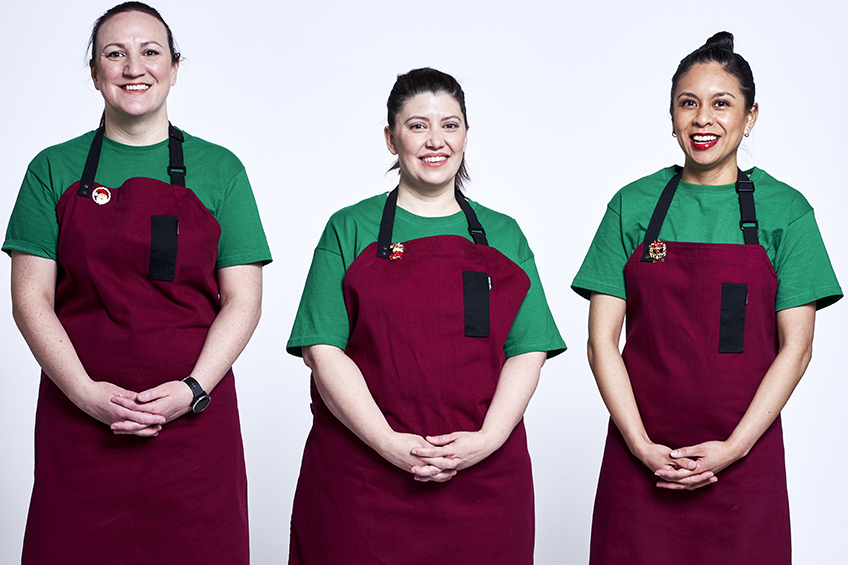 3 contestants looking at the camera and smiling wearing burgundy aprons