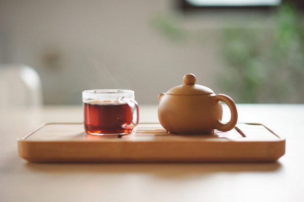 A clear mug of tea on a tray next to a clay tea pot