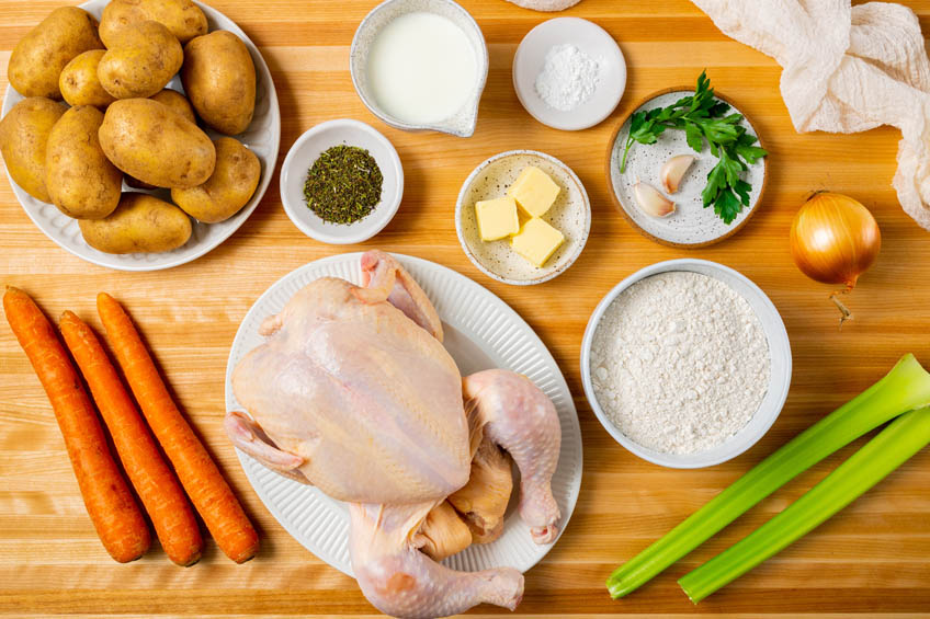 A flat lay of a whole chicken, carrots, celery, onion, potatoes and various spices and herbs