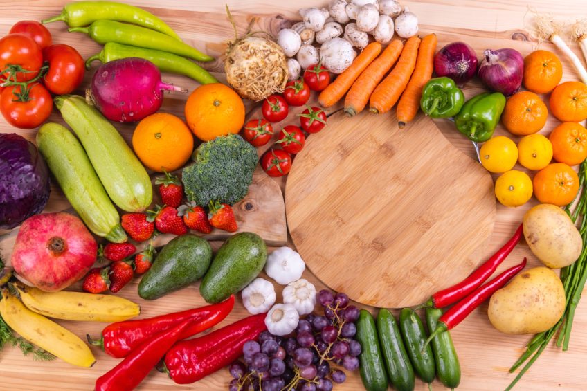 An assortment of fresh fruits and vegetables arranged around a wooden cutting board