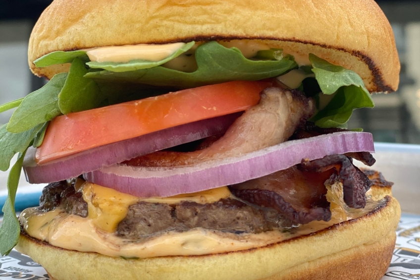 A burger with bacon, Cheddar cheese, arugula, tomato, red onion and sriracha mayo.