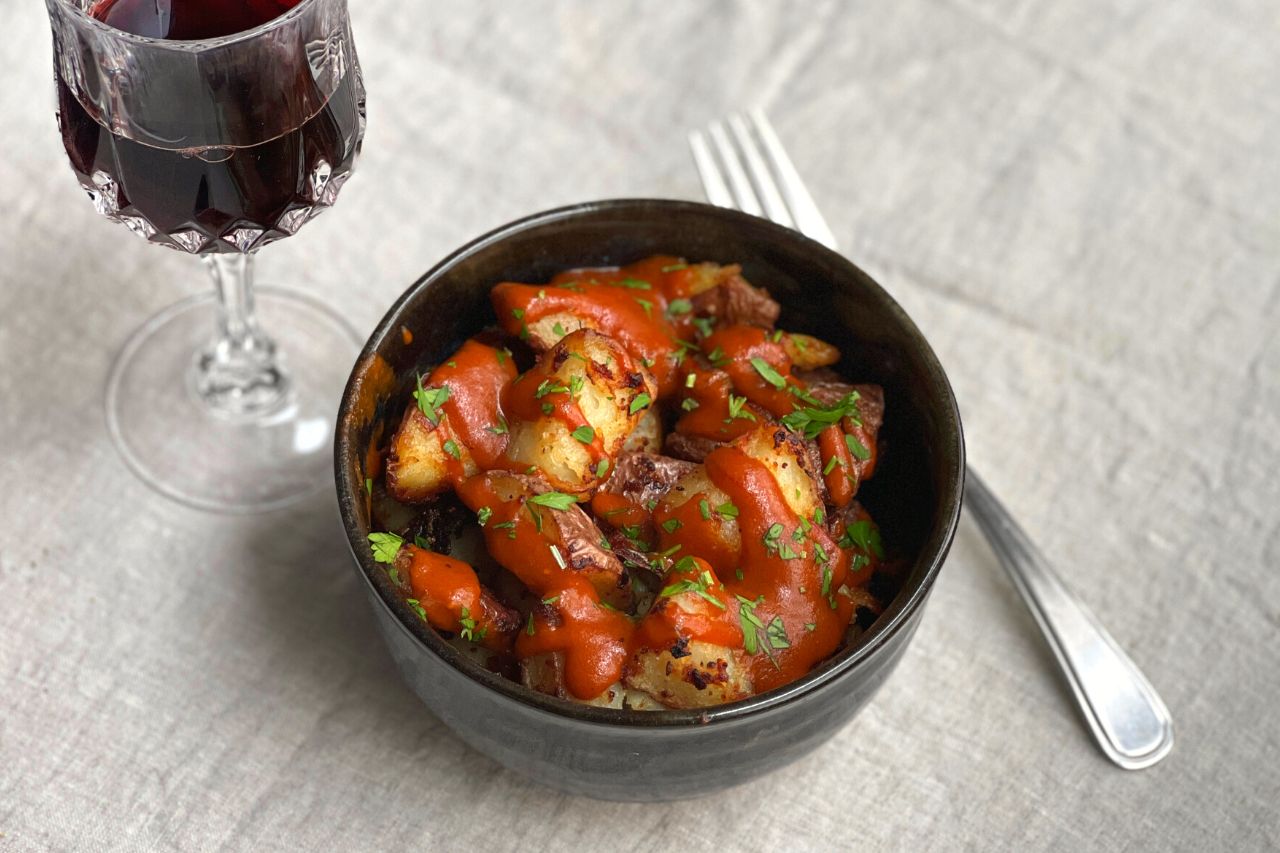 A shot of patatas bravas in a black bowl with a glass of red wine