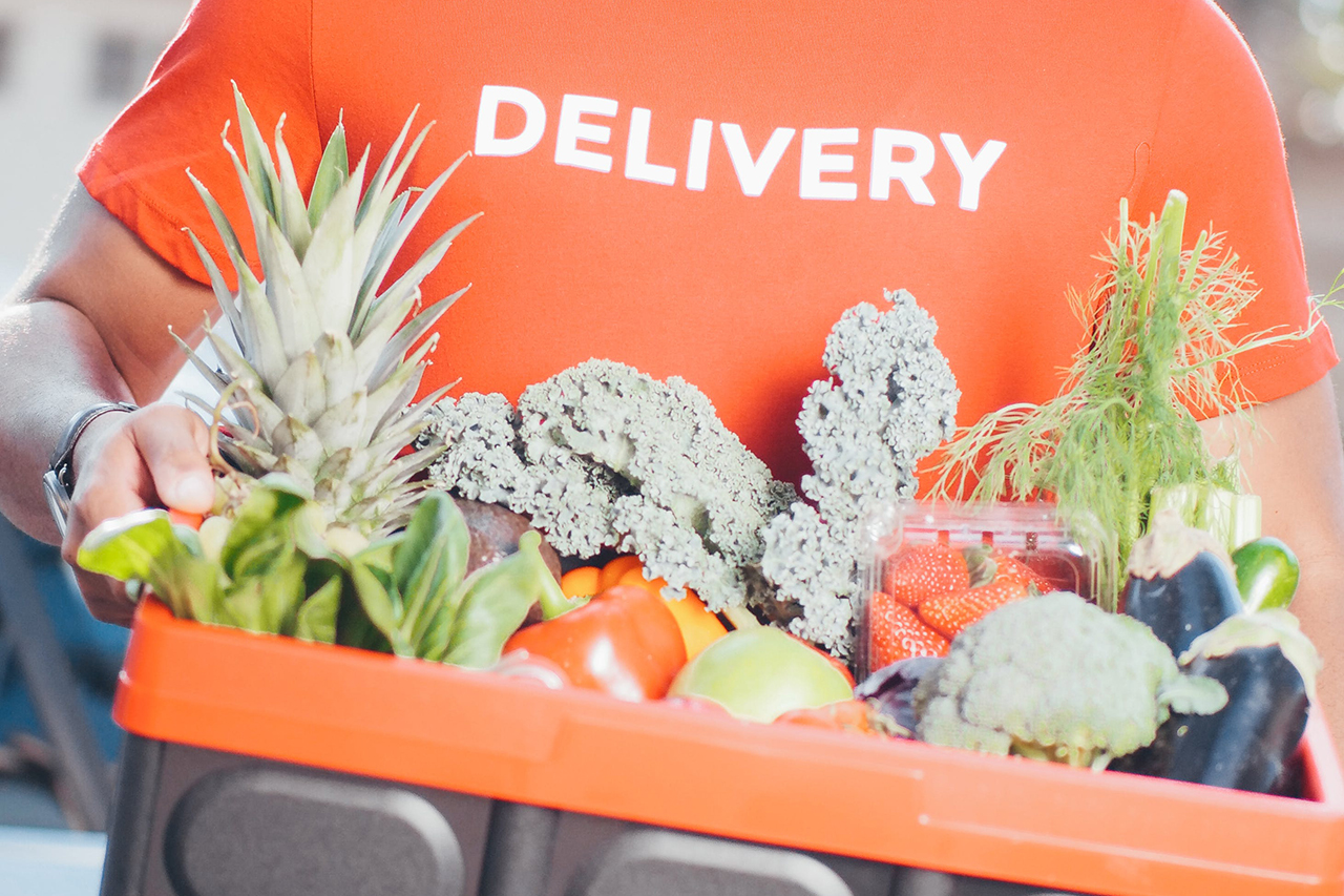 Man wearing a red shirt with the words "delivery" written on them and holding a foldable plastic basket filled with fresh produce.