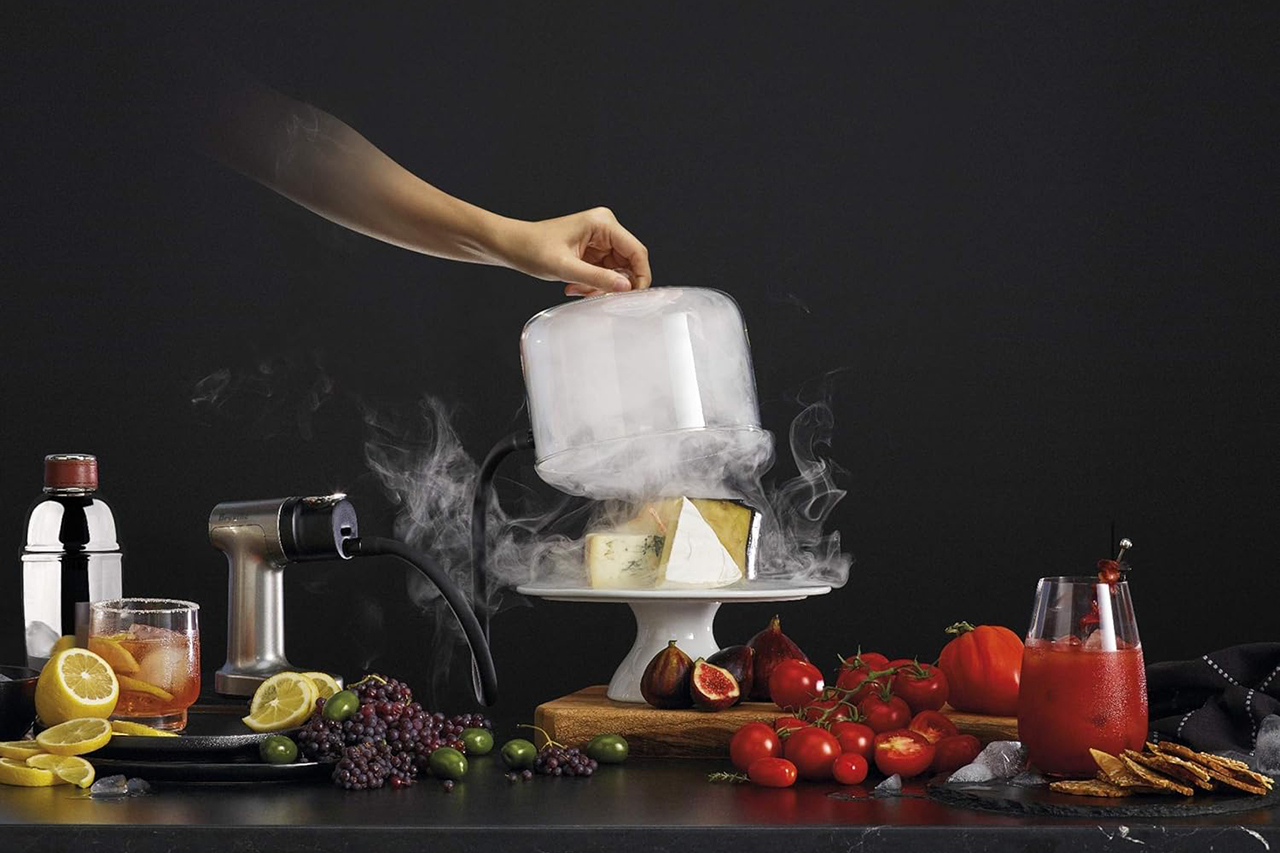 Person lifting a cloche to reveal smoke seeping from underneath on a table full of food