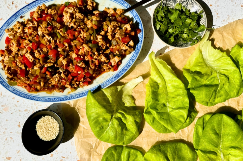 Spicy chicken filling on a plate alongside lettuce leaves, cilantro and sesame seeds